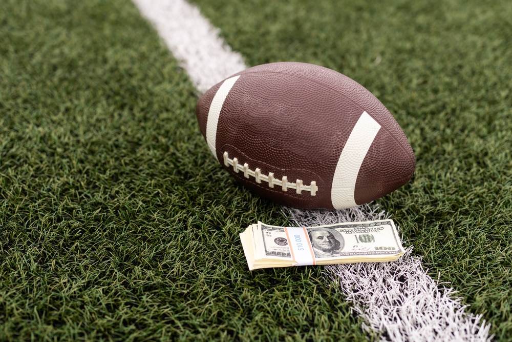A football with a stack of $100 bills sitting next to it on a football field. 